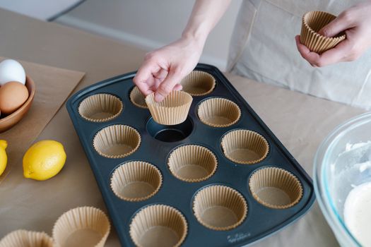 Main plaçant des caissettes en papier dans un moule à cupcakes, avec des ingrédients en arrière-plan.