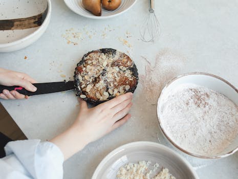 Une main tenant un gâteau au chocolat et poire sur une table de cuisine avec des ingrédients et ustensiles autour.