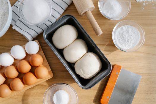 Ingrédients pour brioche maison : pâtes levées, œufs, sucre, farine et ustensiles de cuisine sur une table en bois.