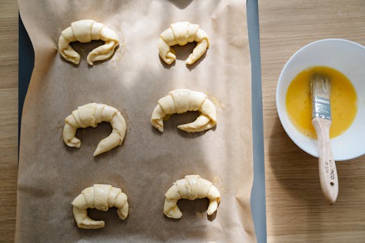 Croissants en pâte feuilletée crue sur une plaque de cuisson, avec un bol d'œuf battu et un pinceau à pâtisserie.
